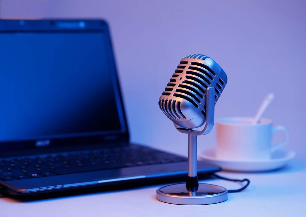 Na ilustração, um microfone em uma mesa ao lado de uma xícara de café e um notebook em alusão ao podcast